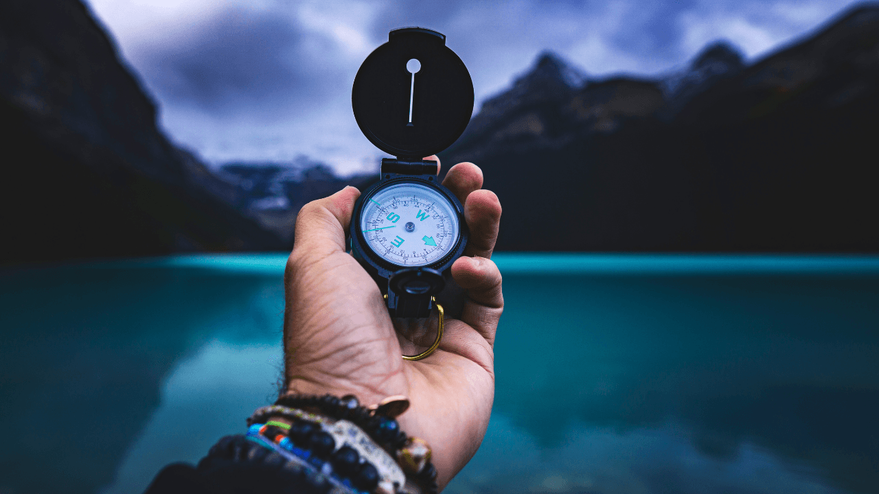 Hand holding a black compass against a backdrop of turquoise water and dark mountains.